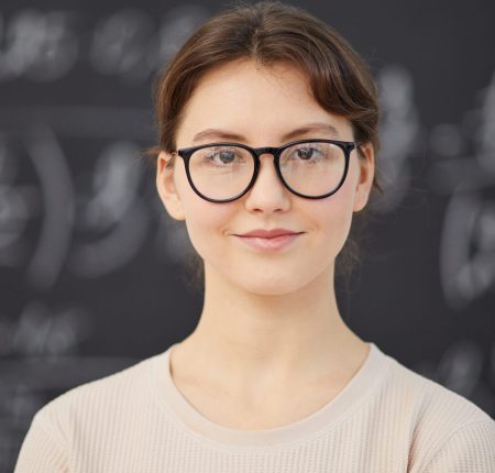 Portrait of female maths teacher in eyeglasses looking at camera with blackboard in the background